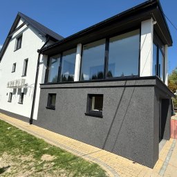 VADYM HAL DOBRO W DZIAŁANIU - Dwukondygnacyjny dom z elewacją w odcieniach bieli i grafitu, z dużymi oknami i kostką brukową wokół. Widoczne klimatyzatory na białej ścianie.