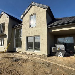 VADYM HAL DOBRO W DZIAŁANIU - Surowa elewacja dwupiętrowego domu z ciemną dachówką, widoczne materiały budowlane i żółty wąż, sugerujące prace wykończeniowe. Błękitne niebo w tle.