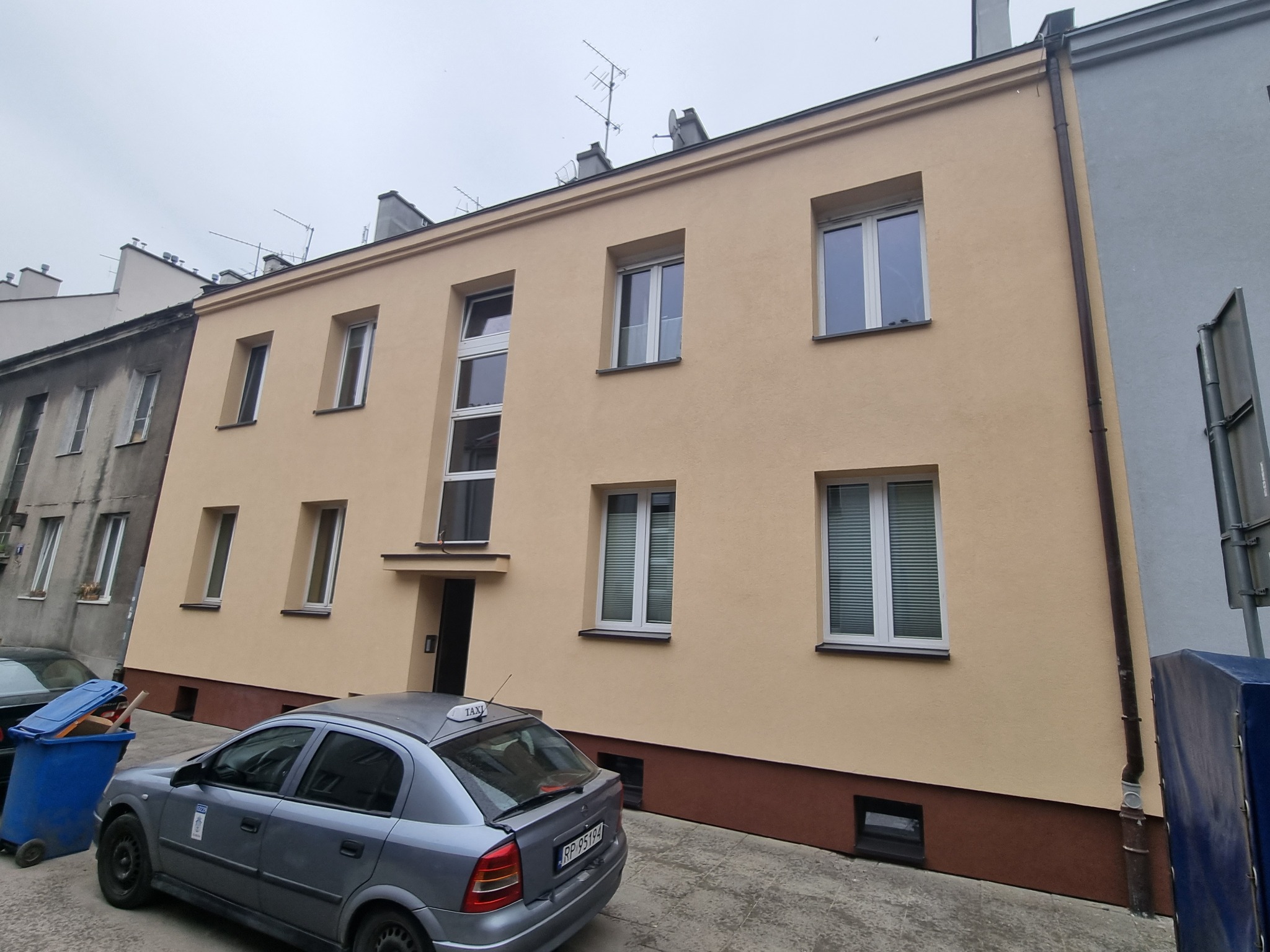 Odnowiona elewacja budynku mieszkalnego w odcieniach beżu i brązu, z widocznym szarym samochodem taxi i niebieskim koszem na śmieci na pierwszym planie.
