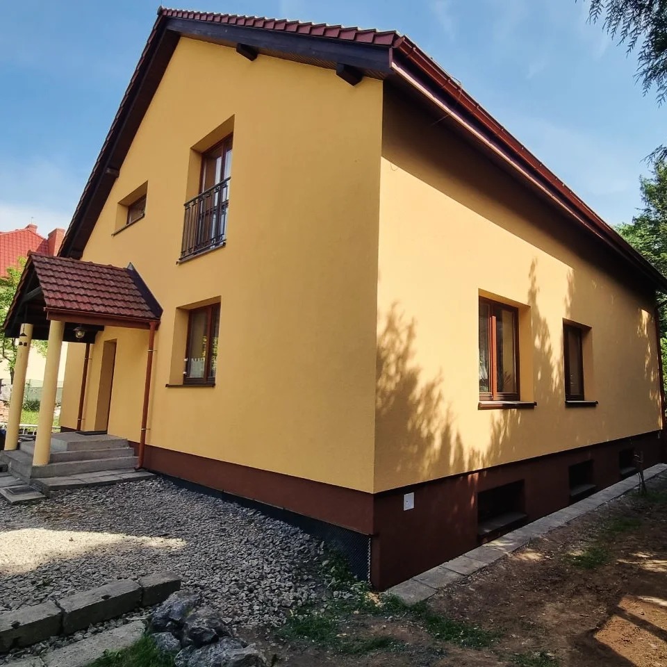 Żółta elewacja domu jednorodzinnego z brązowymi detalami, widok z boku, zadaszone wejście z kolumnami, kamienny podjazd, błękitne niebo w tle.