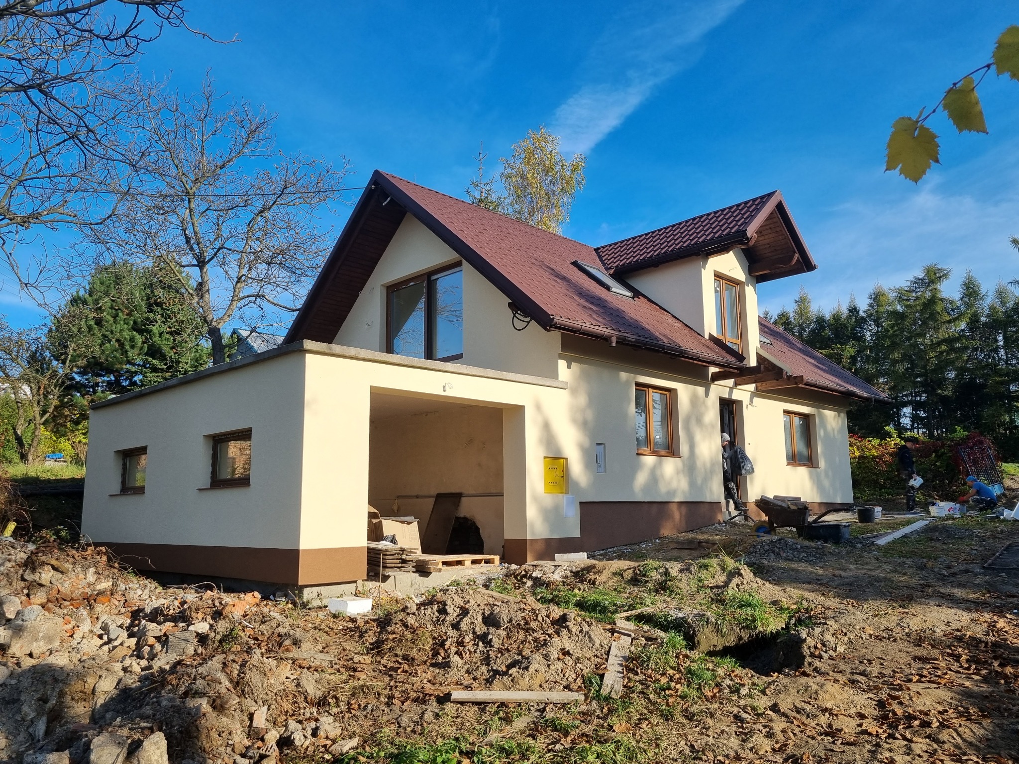 Dwukondygnacyjny dom z brązowym dachem i beżową elewacją, widok z zewnątrz na plac budowy z pracownikami przed wejściem, w tle błękitne niebo