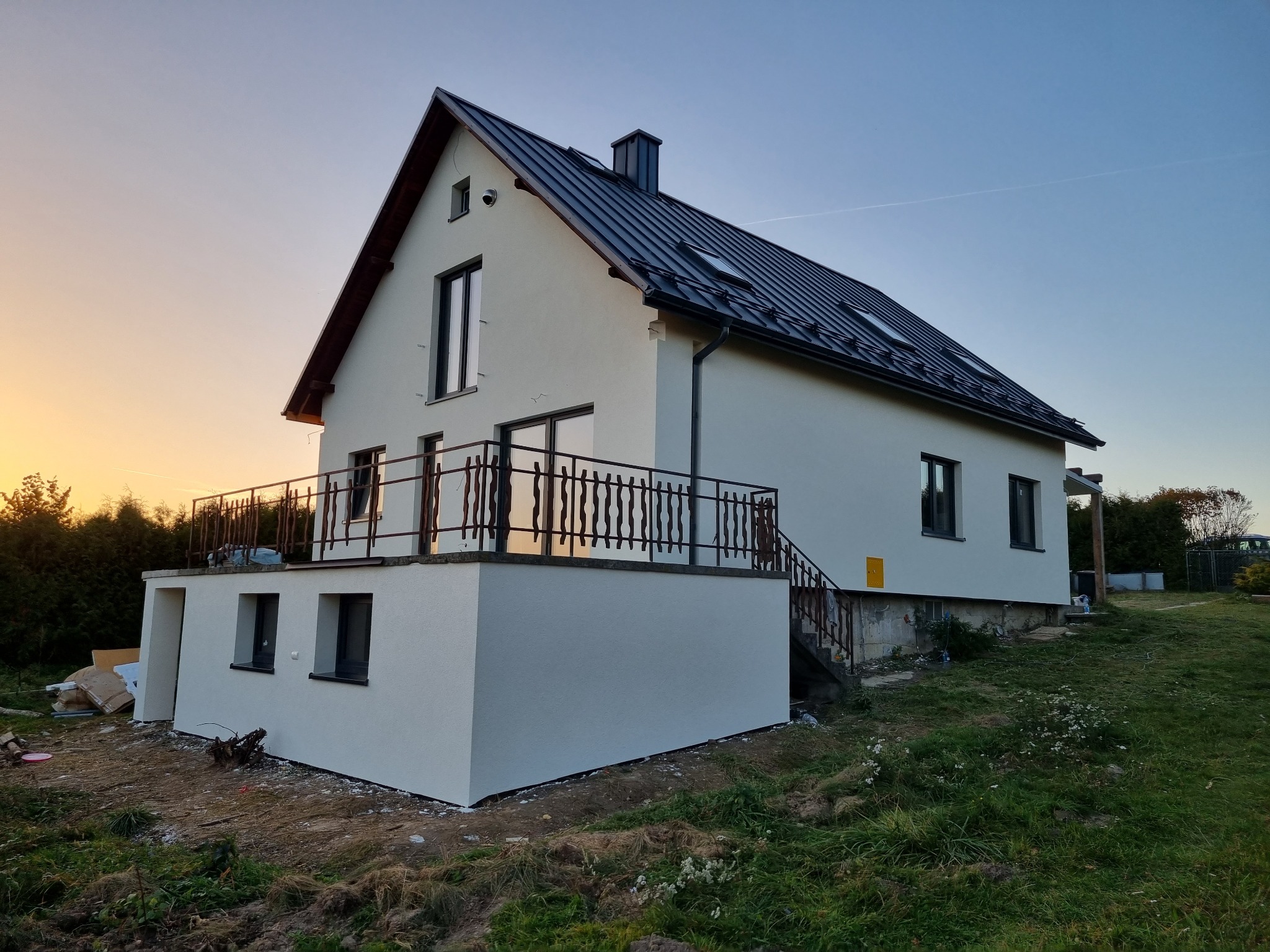 Nowoczesny dom z jasną elewacją i balkonem z ozdobną balustradą, widoczny o zmierzchu na tle zieleni.