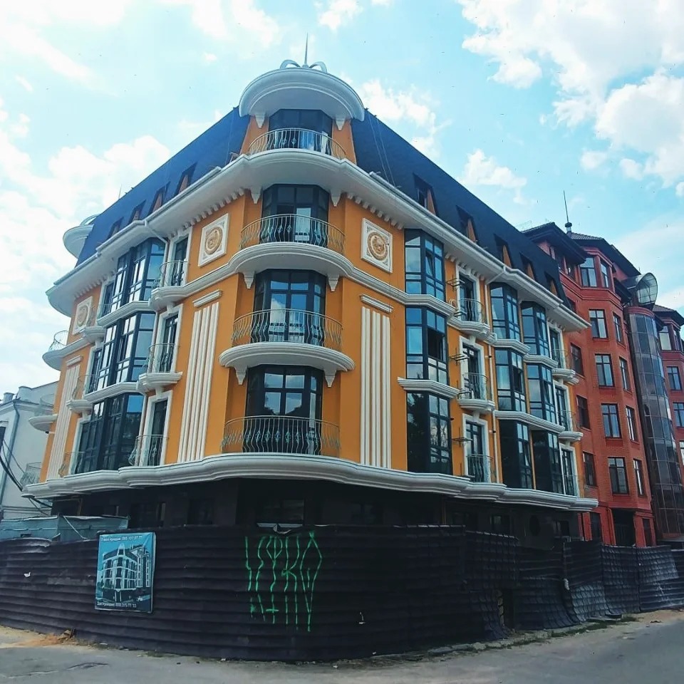 Odnowiony budynek wielorodzinny z pomarańczową elewacją, białymi zdobieniami i balkonami, widoczny spod kąta, z tymczasowym ogrodzeniem i plakatem budowlanym w tle.