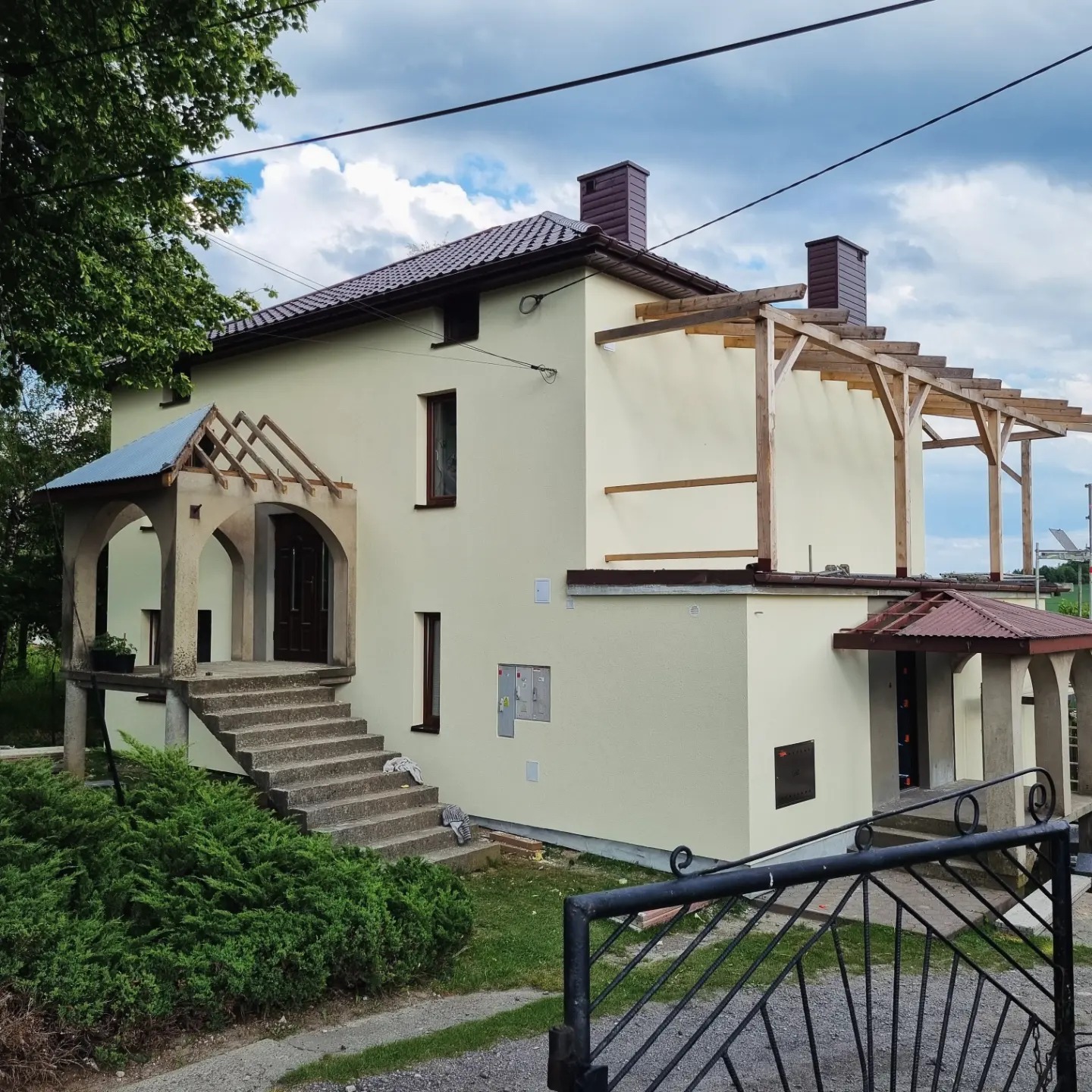 Dwupiętrowy dom z jasną elewacją, drewnianą konstrukcją pergoli i zadaszeniem nad wejściem, widoczne schody wejściowe i metalowe ogrodzenie.