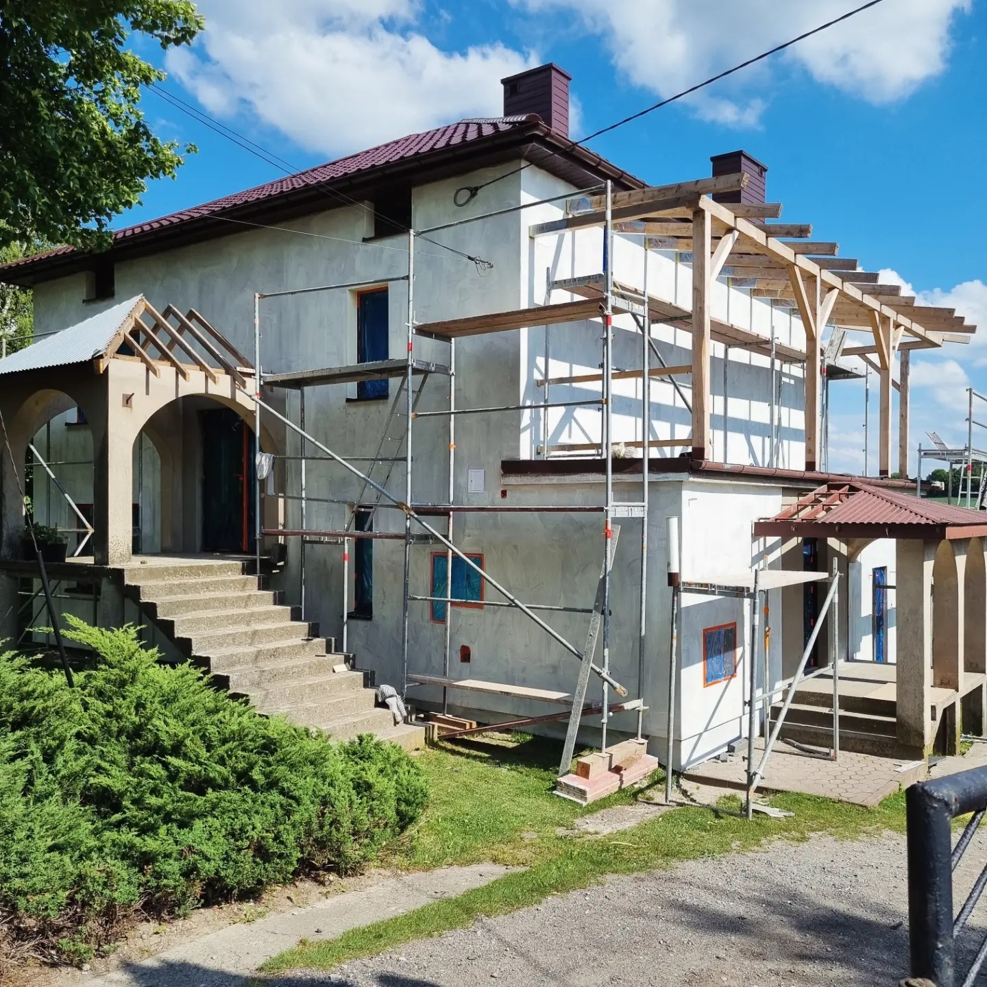 Dom jednorodzinny w trakcie prac elewacyjnych: szare, surowe tynki, rusztowania, drewniana konstrukcja zadaszenia, niebieskie ramy okienne, widok z poziomu gruntu w słoneczny dzień.