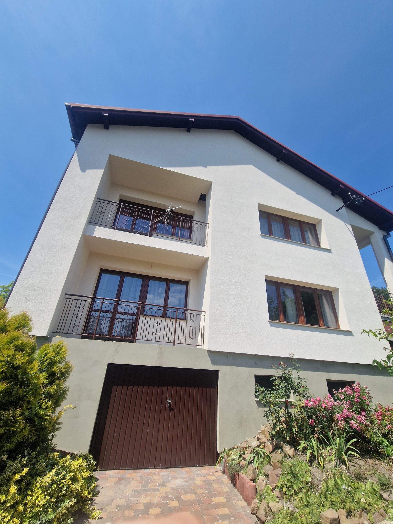 Dwupiętrowy dom z balkonami i garażem, świeżo otynkowany na biało, z ciemnobrązowymi ramami okiennymi i drzwiami garażowymi, widok z dołu na tle błękitnego nieba.