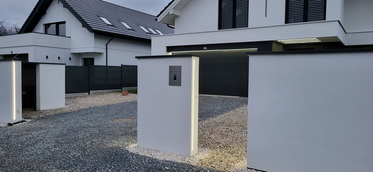 Nowoczesna elewacja domu jednorodzinnego z białym tynkiem, oświetlonymi słupkami ogrodzeniowymi i szarą bramą garażową.