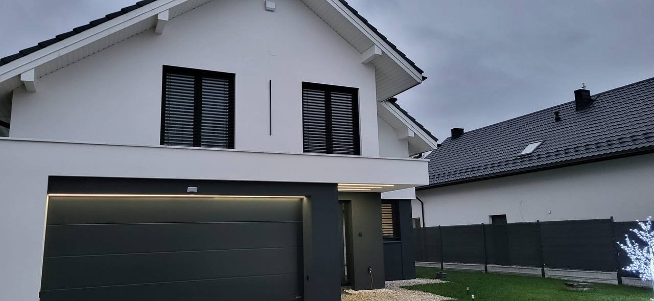 Nowoczesny dom jednorodzinny z białą elewacją, czarnymi oknami z żaluzjami i grafitową bramą garażową z oświetleniem LED, widok z zewnątrz.