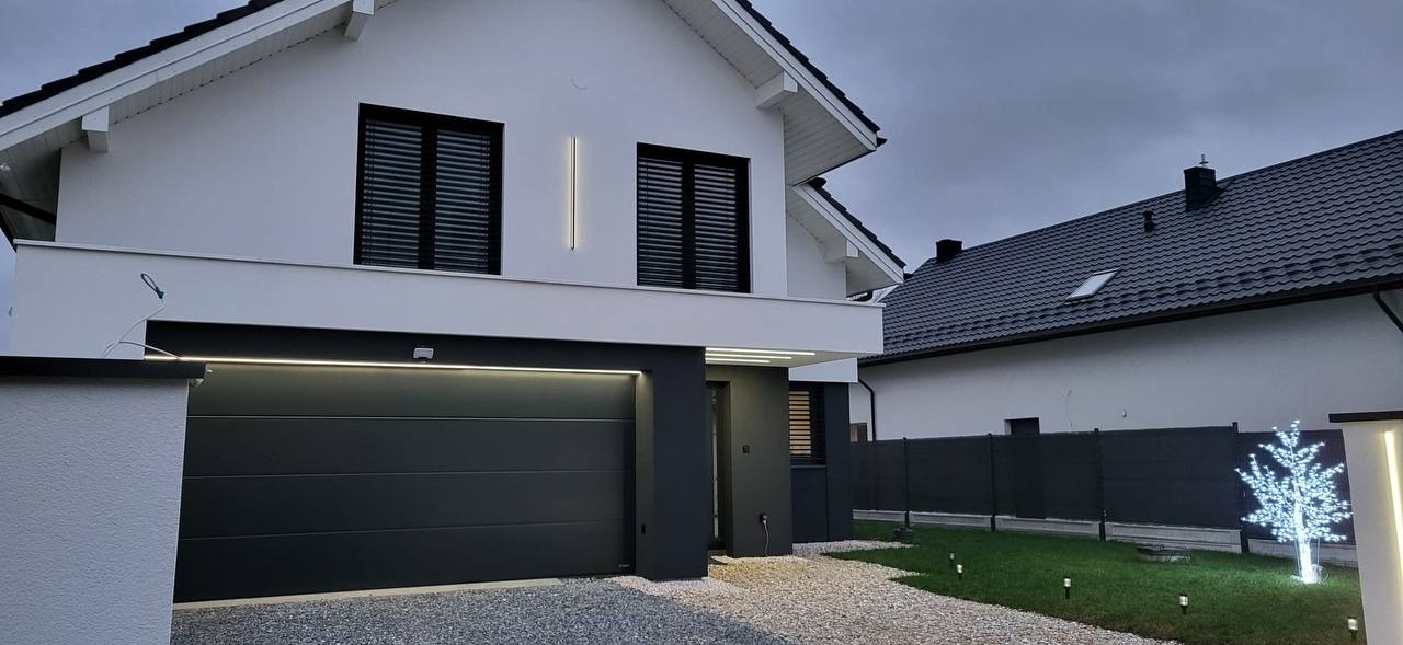 Nowoczesny dom jednorodzinny z białą elewacją, czarnymi oknami z żaluzjami i podświetlanym garażem, oświetlenie elewacyjne w postaci pionowych pasków światła, dekoracyjne drzewko świetlne w ogrodzie.