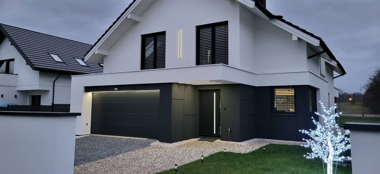 Nowoczesny dom jednorodzinny z szarą elewacją, oświetleniem LED przy oknach i drzwiach, oraz dekoracyjnym drzewkiem świetlnym w ogrodzie.