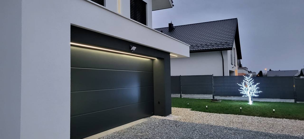 Nowoczesna, grafitowa brama garażowa segmentowa z poziomymi przetłoczeniami, oświetlona listwą LED, wkomponowana w elewację domu z oświetlonym drzewkiem dekoracyjnym w ogrodzie.