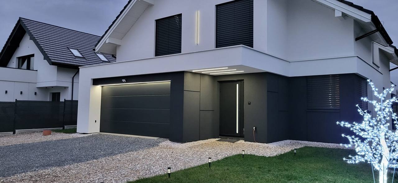 Nowoczesny dom jednorodzinny z grafitową elewacją, oświetleniem LED przy drzwiach wejściowych i bramie garażowej, oraz dekoracyjnym drzewkiem świetlnym na trawniku.