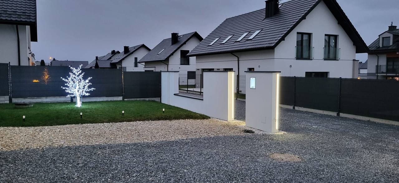 Nowoczesne osiedle domów jednorodzinnych z białymi elewacjami, czarnymi dachami i grafitowymi ogrodzeniami, oświetlone dekoracyjnymi lampami i drzewkiem LED.