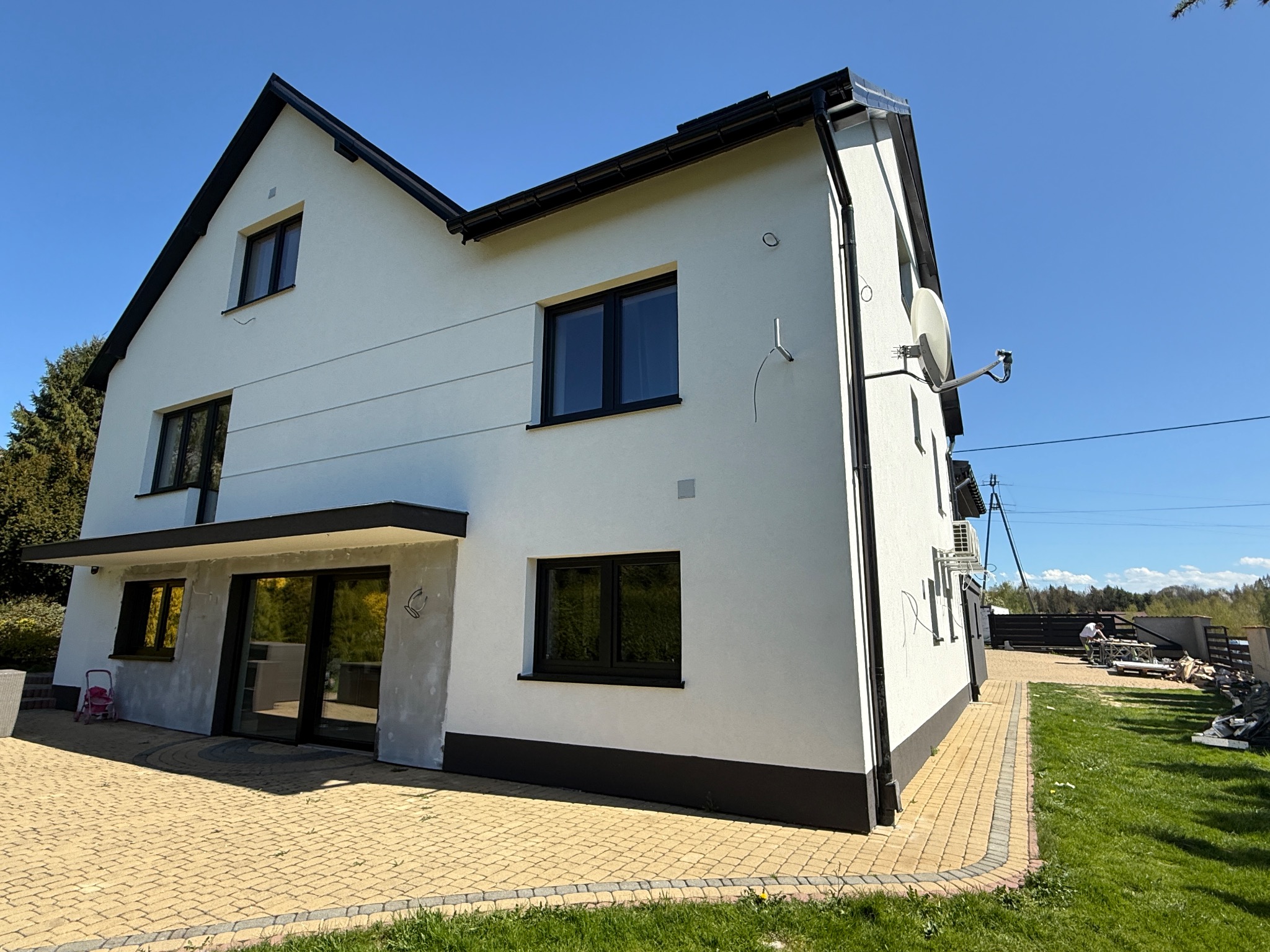 Dwukondygnacyjny dom z białą elewacją i czarnymi ramami okiennymi, widziany z perspektywy ogrodu z kostką brukową. Świeżo pomalowana fasada w słoneczny dzień.