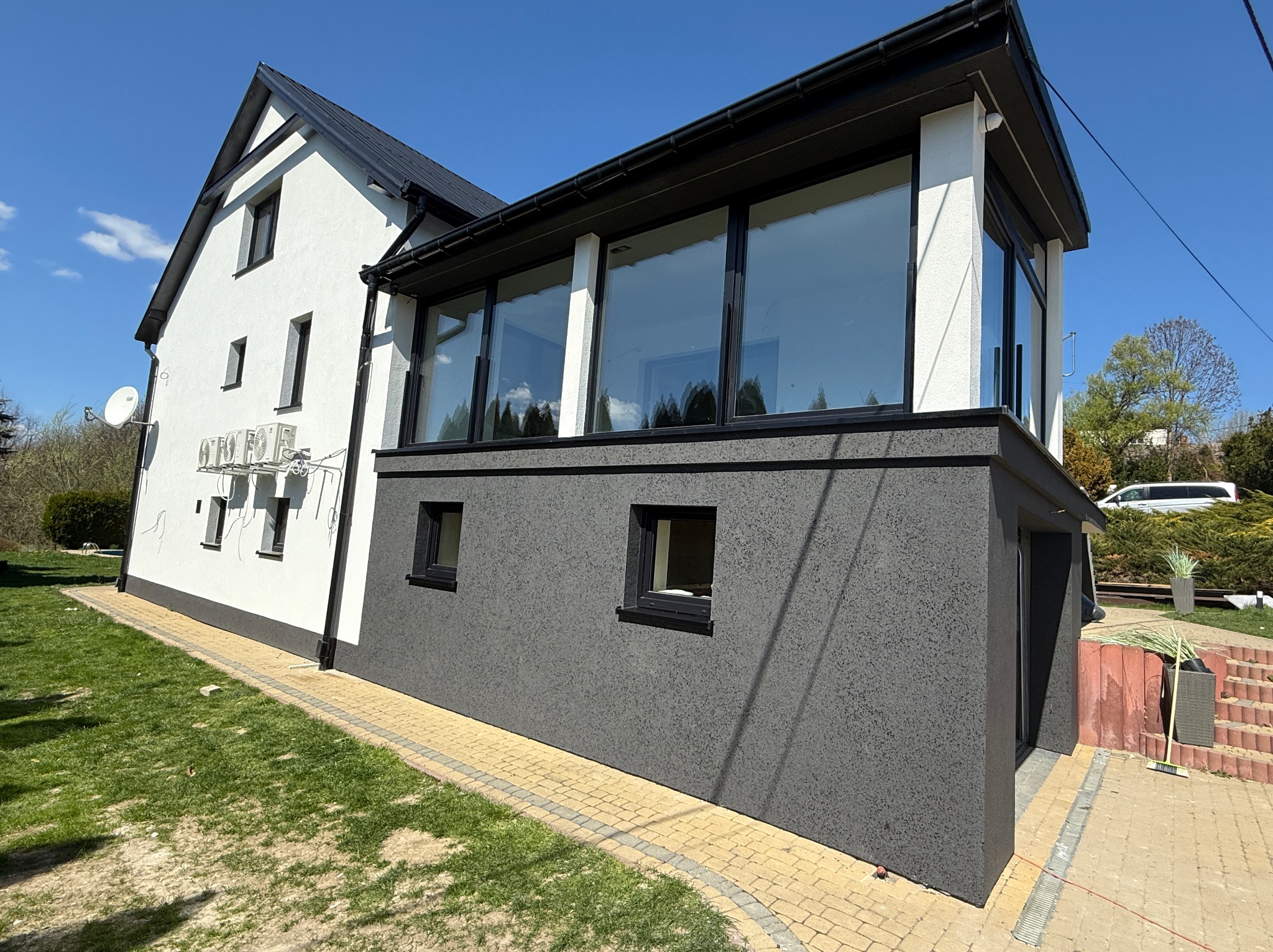 Dwukondygnacyjny dom z elewacją w odcieniach bieli i grafitu, z dużymi oknami i kostką brukową wokół. Widoczne klimatyzatory na białej ścianie.