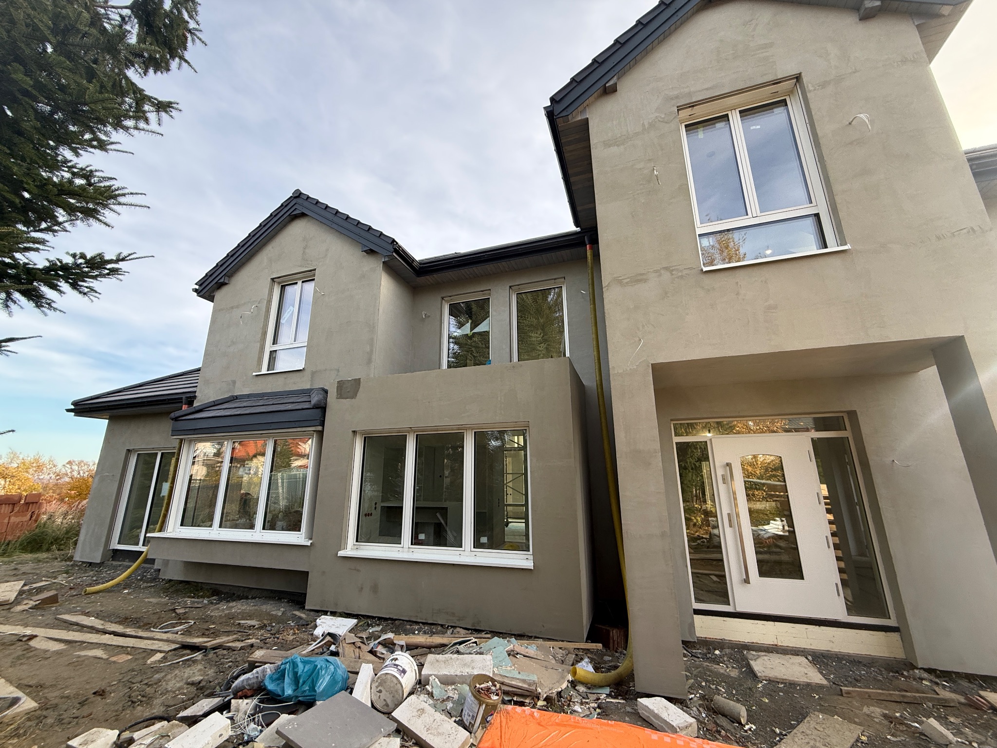 Dom dwupiętrowy w trakcie tynkowania elewacji zewnętrznej, widoczne białe okna i drzwi wejściowe. Na ziemi gruz i materiały budowlane. Szary odcień tynku.