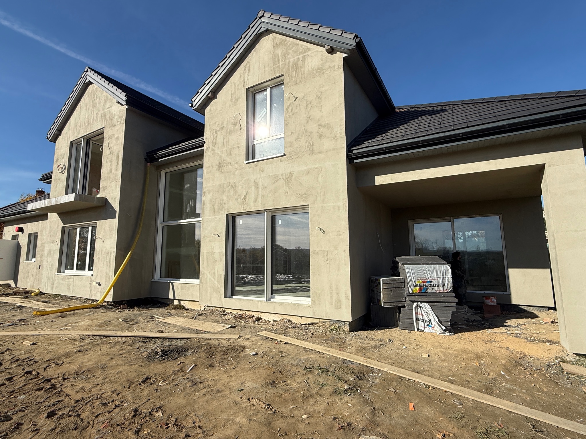 Surowa elewacja dwupiętrowego domu z ciemną dachówką, widoczne materiały budowlane i żółty wąż, sugerujące prace wykończeniowe. Błękitne niebo w tle.