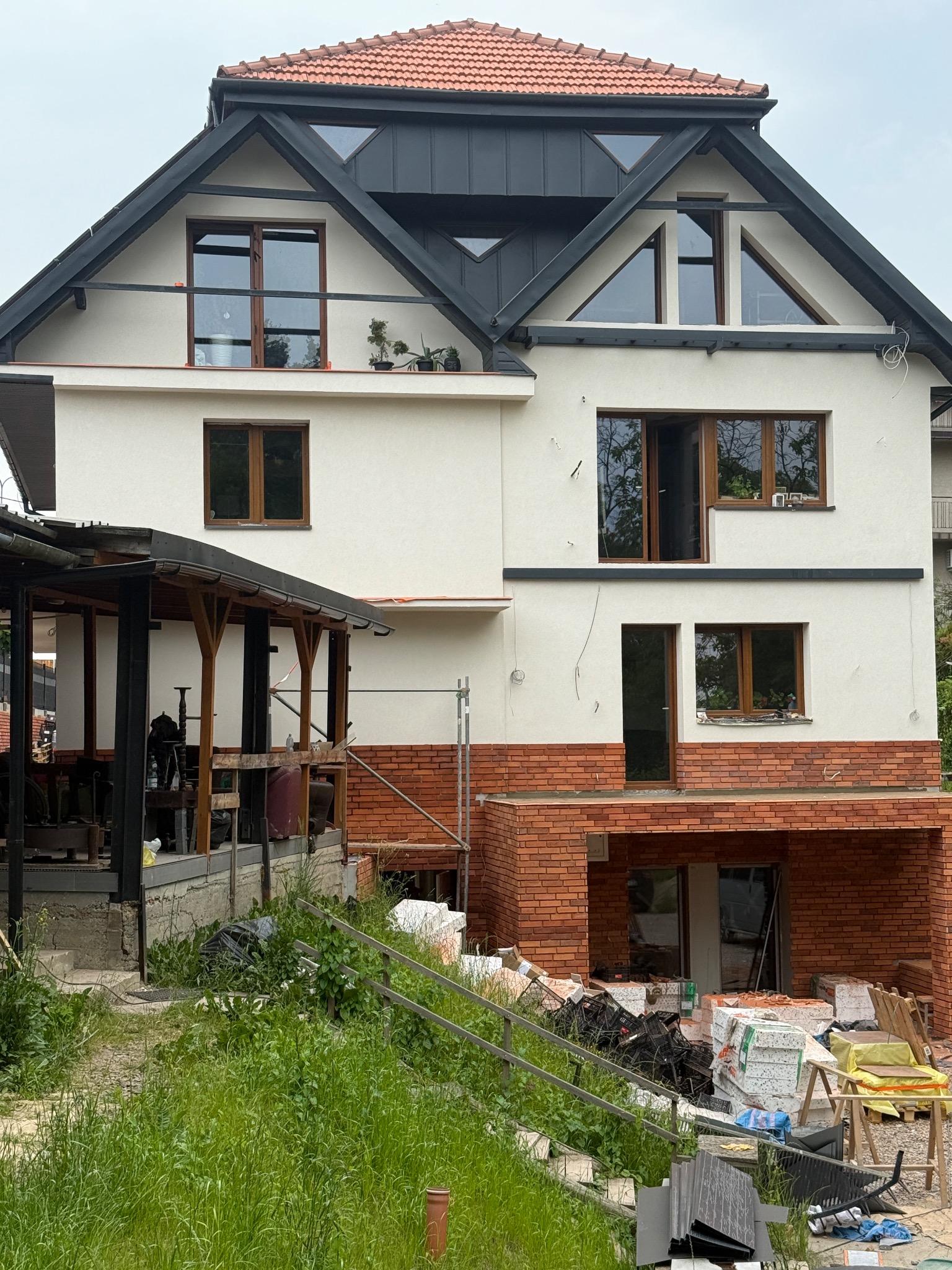 Dom w trakcie prac elewacyjnych: biały tynk, ceglana podmurówka, drewniane okna. Widoczne materiały budowlane i rusztowania. Dach z czerwoną dachówką.