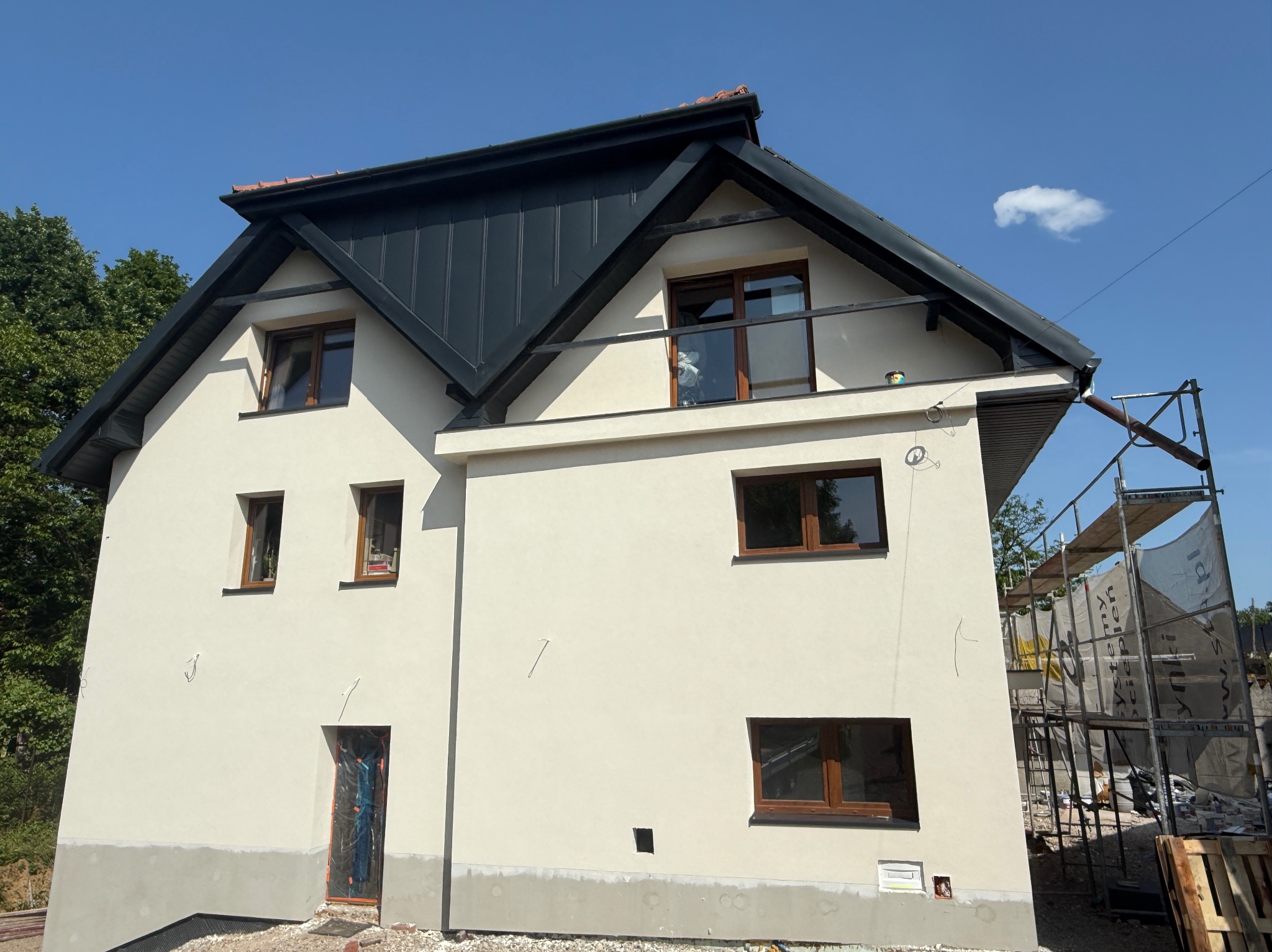Dwupiętrowy dom z jasną elewacją i ciemnym dachem, okna z brązowymi ramami. Częściowo widoczne rusztowania. Błękitne niebo z pojedynczą chmurą w tle.