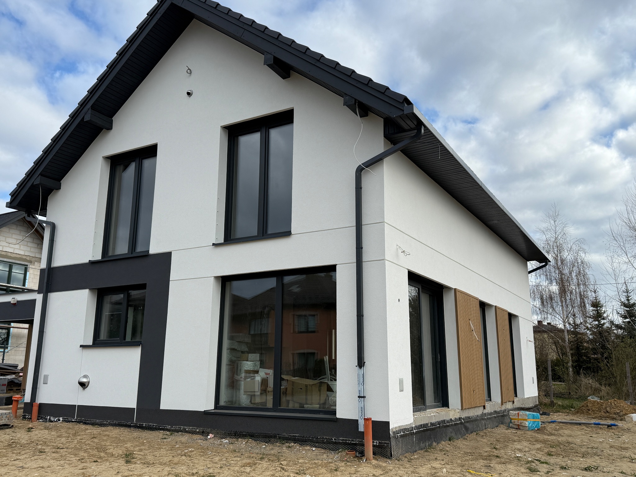 Nowoczesny dom jednorodzinny z białą elewacją i czarnymi akcentami, duże okna, częściowo wykończona elewacja, widoczne materiały budowlane na posesji.
