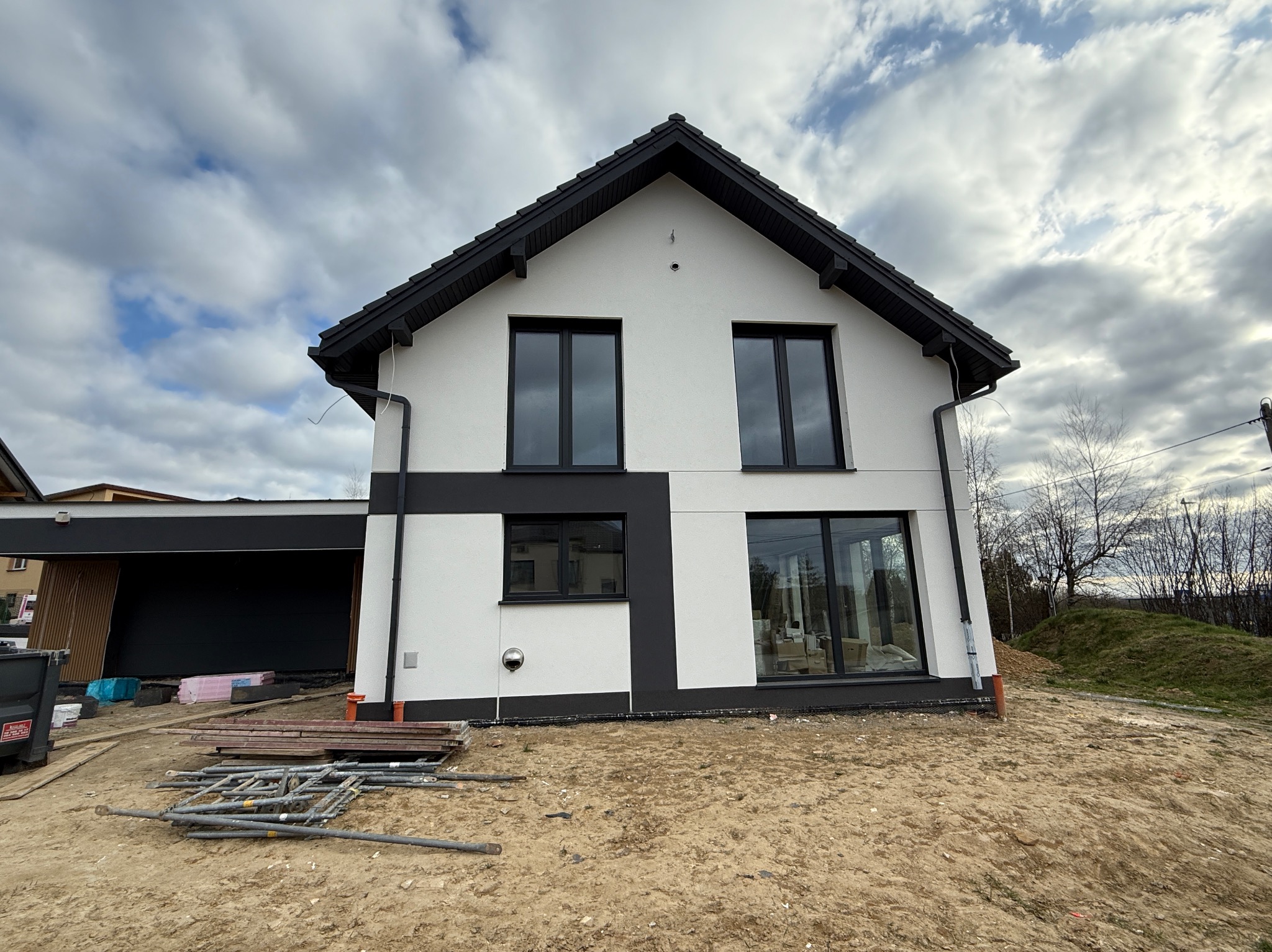 Nowoczesny dom jednorodzinny z białą elewacją i czarnymi akcentami, widok z zewnątrz, teren wokół domu w trakcie prac wykończeniowych, pochmurne niebo.