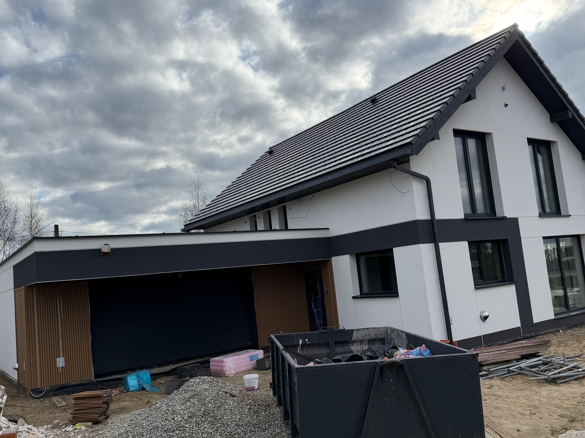 Nowoczesny dom jednorodzinny z grafitowym dachem i białymi ścianami, w trakcie prac wykończeniowych elewacji. Widoczny kontener na gruz i materiały budowlane.