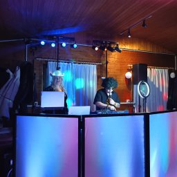 ElDżi&Eva LiveMusik - Dwoje wykonawców za podświetlonym stanowiskiem DJ-skim podczas występu, oświetlenie sceniczne i drewniana boazeria w tle.