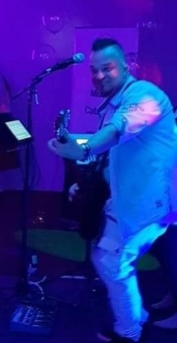 Gitarzysta z irokezem, grający na scenie w fioletowym oświetleniu, z mikrofonem na statywie i pulpitem na nuty obok.