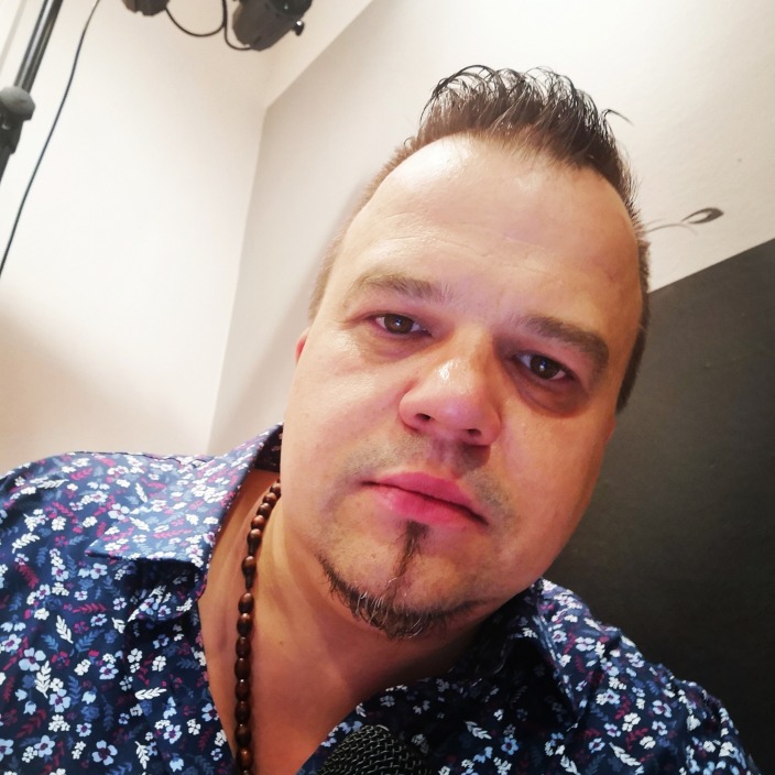 Portret mężczyzny z mikrofonem, w koszuli w kwiaty i z drewnianym naszyjnikiem, patrzącego w obiektyw.