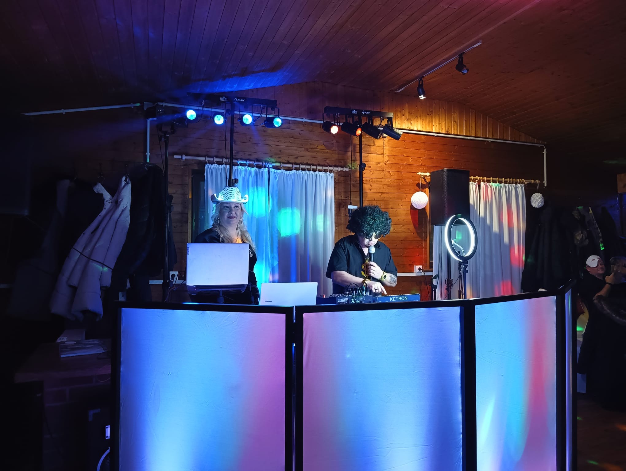 Dwoje wykonawców za podświetlonym stanowiskiem DJ-skim podczas występu, oświetlenie sceniczne i drewniana boazeria w tle.