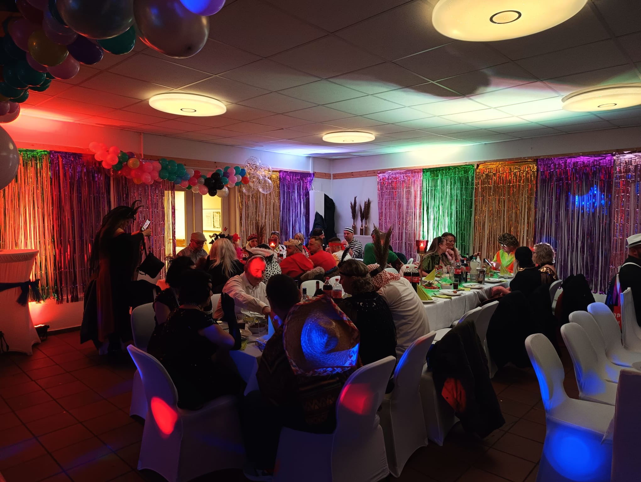 Uczestnicy imprezy kostiumowej siedzą przy udekorowanym stole, oświetlonym kolorowymi światłami i ozdobionym błyszczącymi kurtynami oraz balonami.