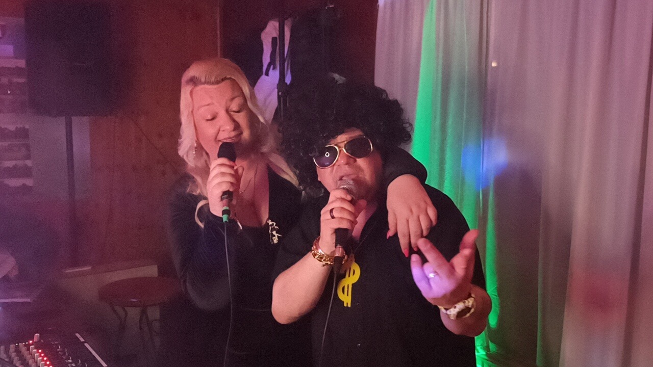 Duet wokalny: kobieta z blond włosami i mężczyzna w afro peruce i okularach przeciwsłonecznych, oboje śpiewają do mikrofonów na tle kurtyny z zielonym oświetleniem.