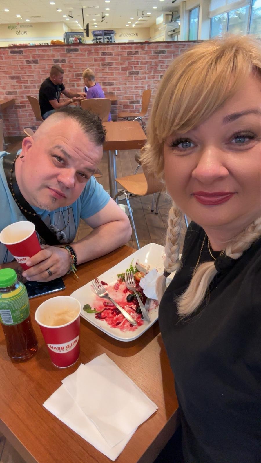 Dwie osoby przy stole w restauracji, po posiłku, widoczny talerz z resztkami sałatki, kubki po napojach i butelka, ceglana ściana w tle.