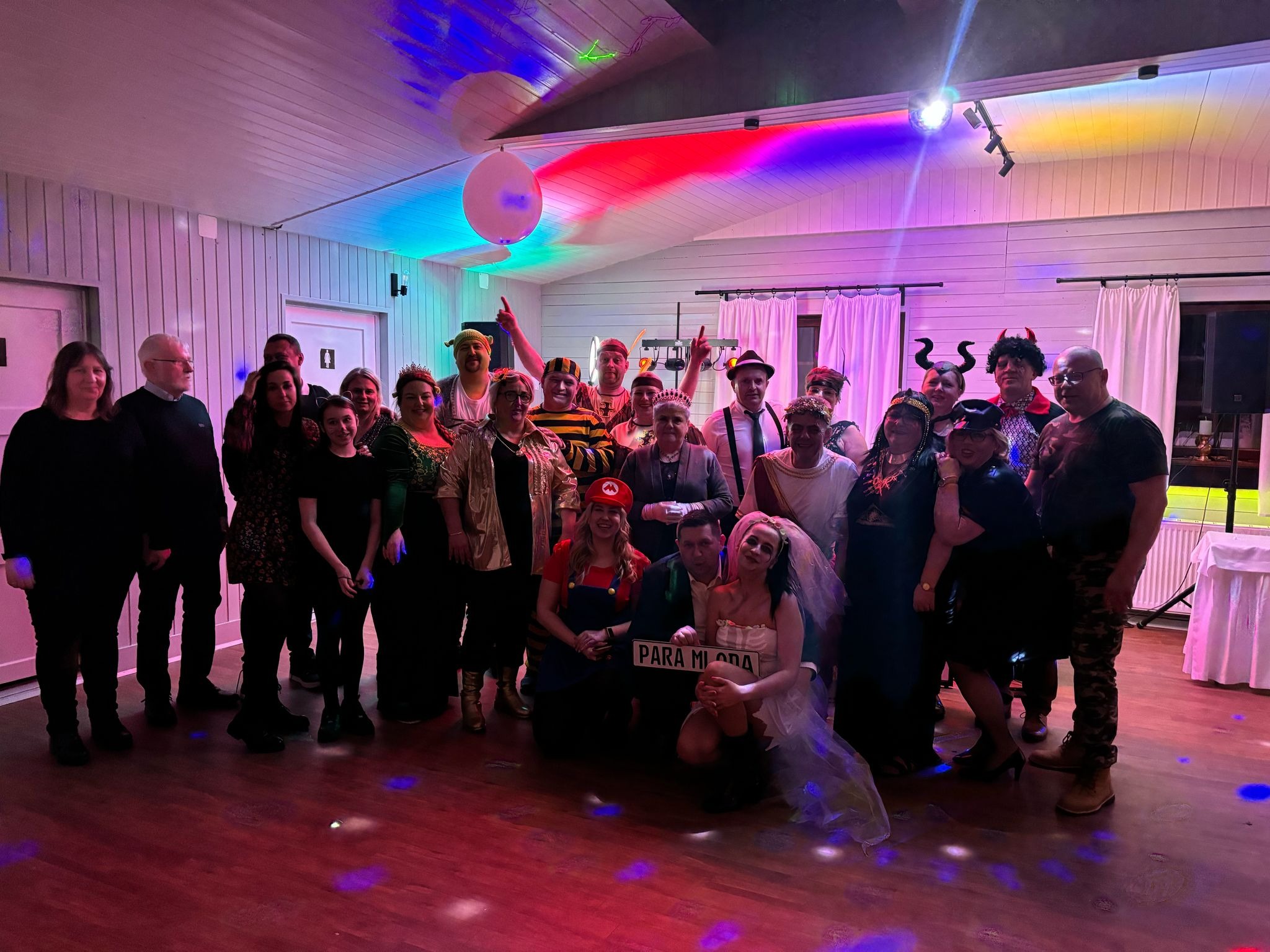 Grupa osób w różnorodnych kostiumach na imprezie, oświetlona kolorowymi światłami, z parą młodą trzymającą tabliczkę 'Para Młoda'.