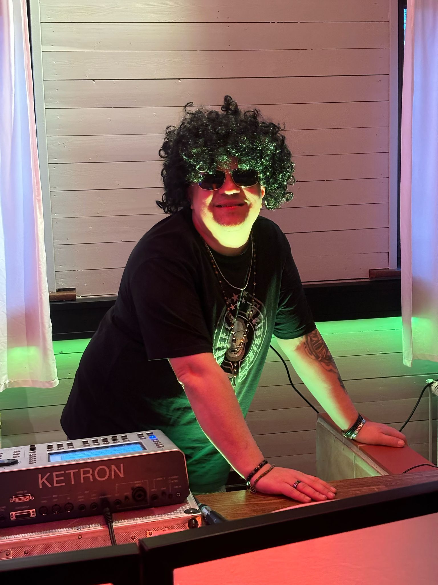 Osoba w czarnej koszulce i peruce afro za konsoletą Ketron, w okularach przeciwsłonecznych, oświetlona kolorowym światłem.