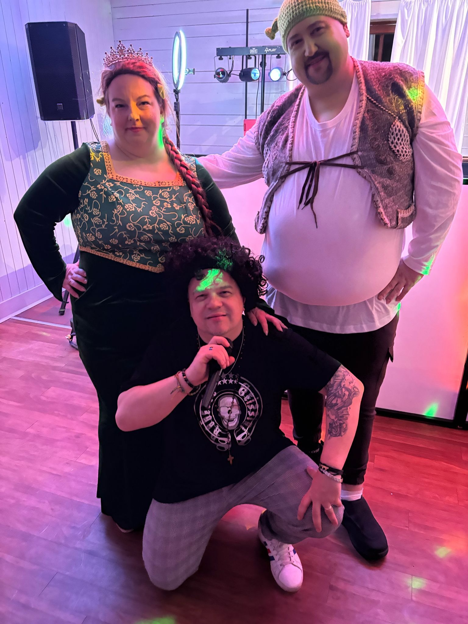 Trzy osoby przebrane za postacie z bajki Shrek: Fiona, Shrek i osioł, pozują do zdjęcia na tle sceny z oświetleniem punktowym.