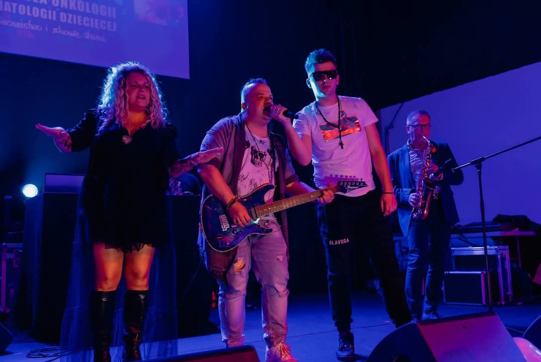 Zespół muzyczny podczas występu na scenie, wokalistka z rozłożonymi rękami, gitarzysta z mikrofonem, mężczyzna w okularach i saksofonista w tle.