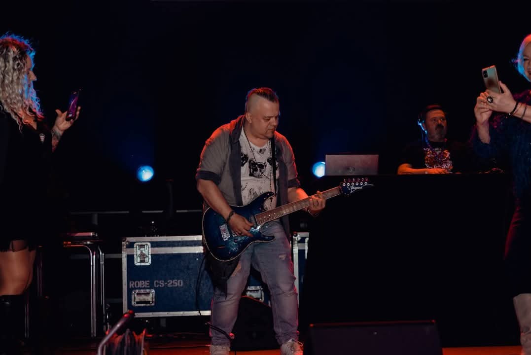 Gitarzysta w jeansach i koszulce z nadrukiem gra na niebieskiej gitarze elektrycznej na scenie podczas koncertu, w tle osoby nagrywające występ telefonami.