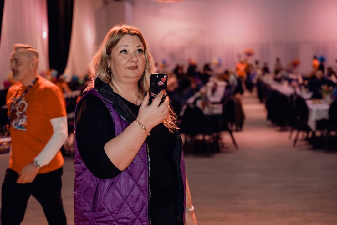 Kobieta w fioletowej kamizelce robi zdjęcie telefonem na tle sali bankietowej z ustawionymi stołami i siedzącymi gośćmi.
