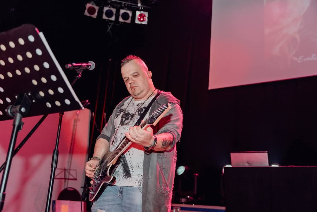 Gitarzysta z irokezem gra na czarnej gitarze elektrycznej na scenie z mikrofonem i pulpitem do nut, w tle czerwone oświetlenie i ekran projekcyjny.