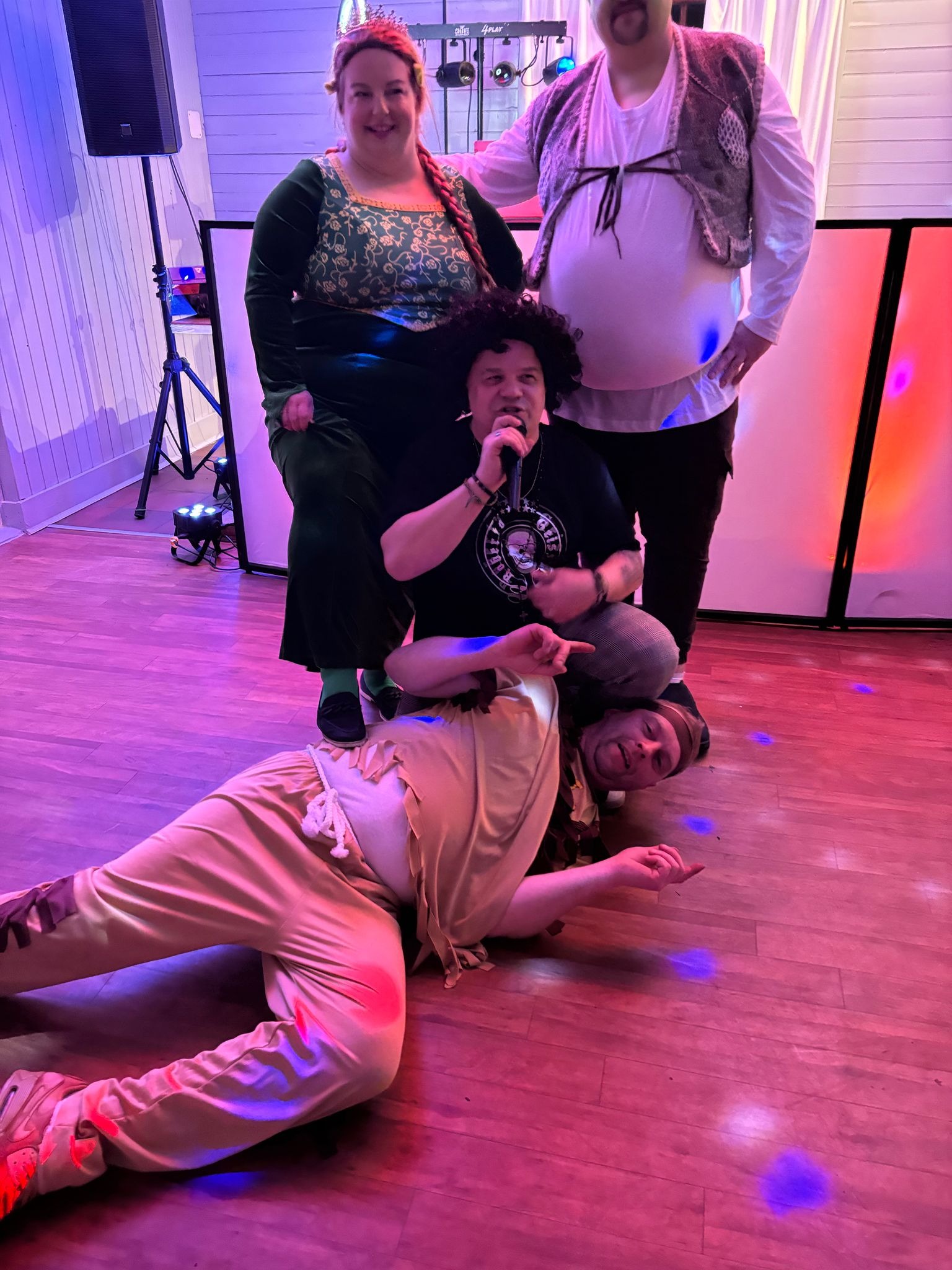 Grupa osób w kostiumach na scenie, w tym osoba z mikrofonem i oświetlenie sceniczne.