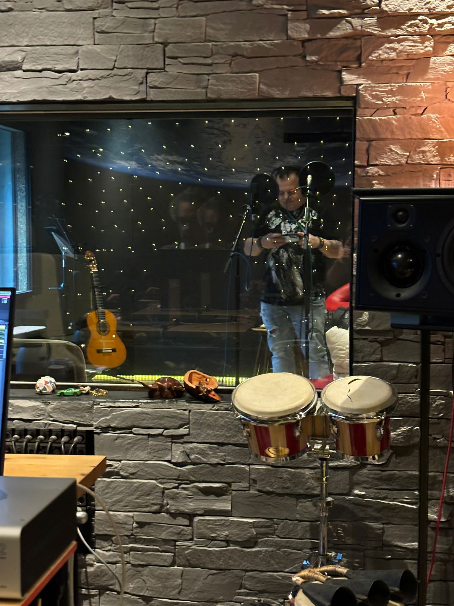 Wnętrze studia nagraniowego z kamienną ścianą, widoczne bębny bonga, głośnik, gitara klasyczna i muzyk za szybą, sprawdzający coś na telefonie, z mikrofonami przed sobą.