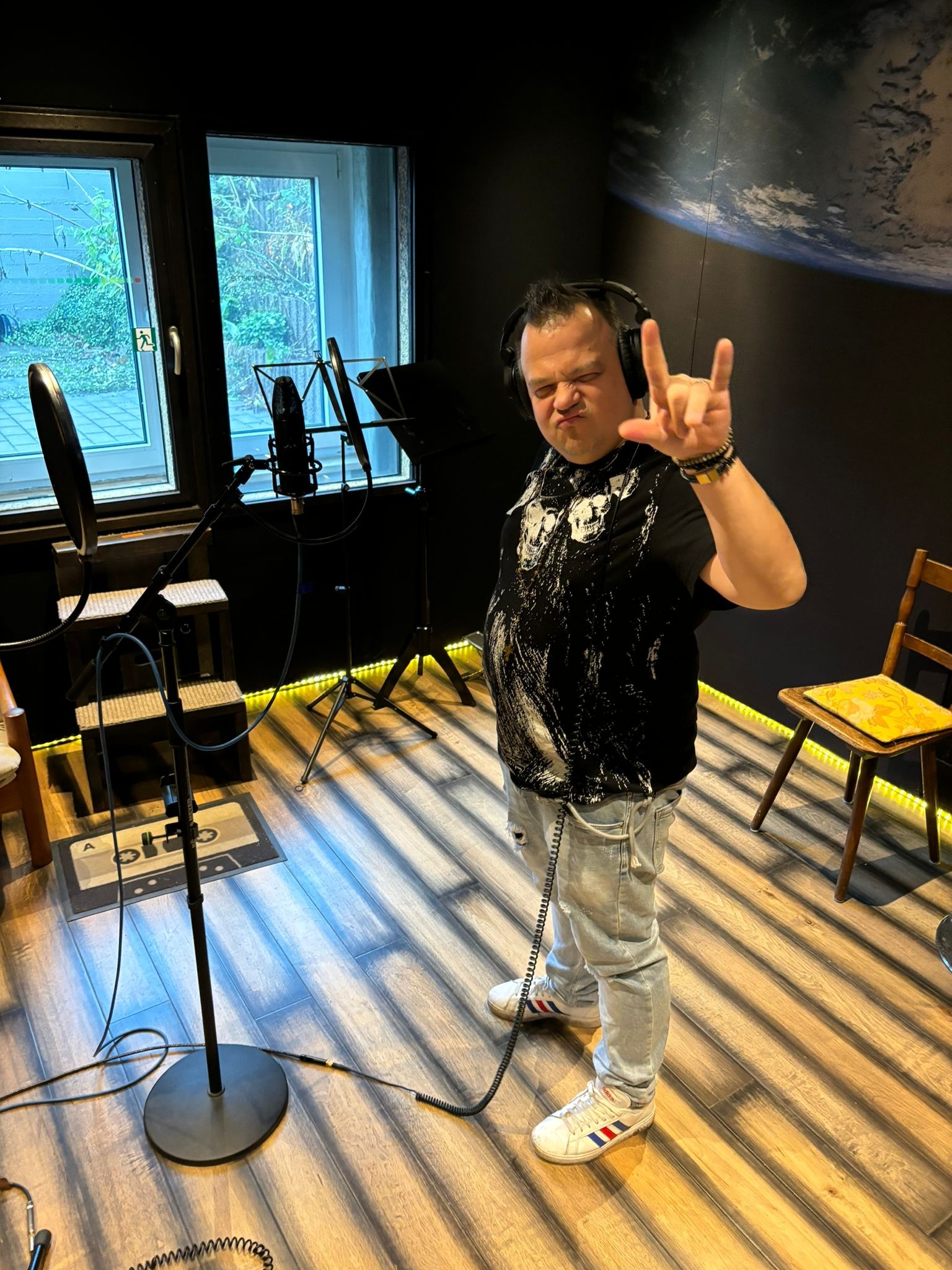 Mężczyzna w słuchawkach, przed mikrofonem studyjnym, wykonuje gest dłońmi na tle ściany z motywem kosmosu w domowym studio nagrań.