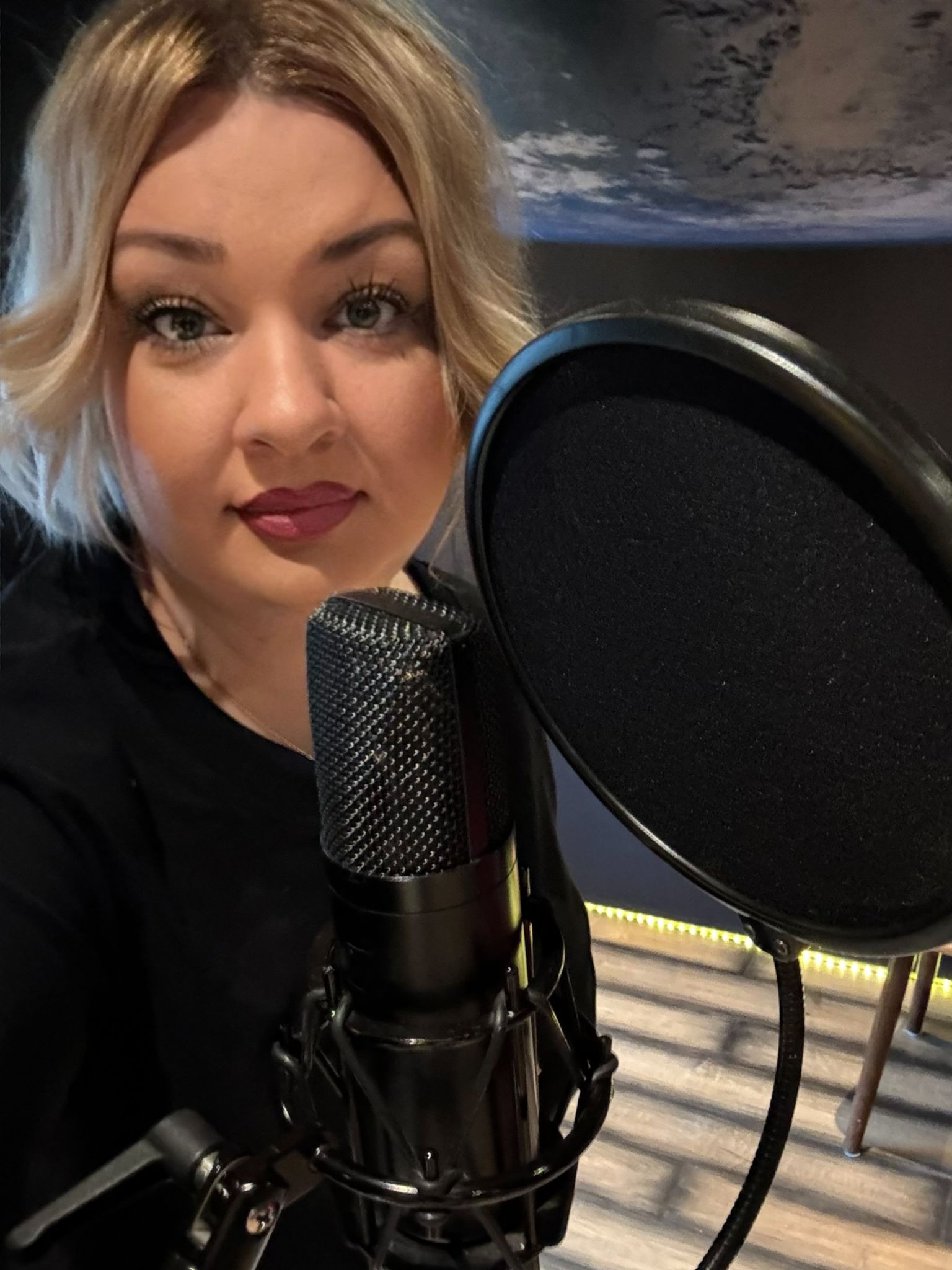 Portret kobiety z blond włosami i mikrofonem studyjnym z filtrem pop, w tle podświetlona podłoga i grafika z motywem kosmicznym.