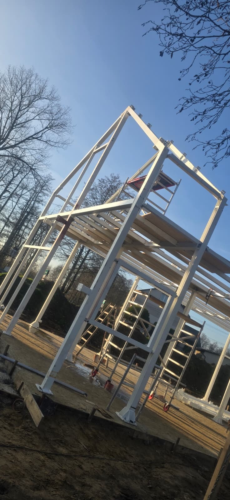 Wznoszenie szkieletu domu z białych, metalowych profili na tle błękitnego nieba i drzew. Widoczne drabiny i elementy konstrukcyjne. Ujęcie z dołu.