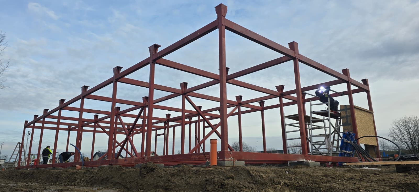 Czerwona konstrukcja stalowa hali w trakcie budowy, spawacz na drabinie, pochmurne niebo w tle. Widoczne elementy zbrojenia i fundamenty.