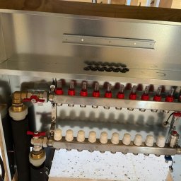 Usługi hydrauliczne Jakub Mejna - Rozdzielacz ogrzewania podłogowego z odpowietrznikami, zamontowany w skrzynce, widoczne przyłącza i izolacja, surowe drewniane elementy konstrukcyjne w tle.