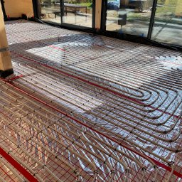 Usługi hydrauliczne Jakub Mejna - Białe rurki ogrzewania podłogowego ułożone na folii izolacyjnej z czerwoną siatką w nowym domu z dużymi oknami.