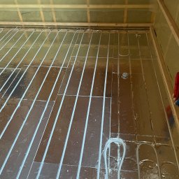 Usługi hydrauliczne Jakub Mejna - Montaż rur ogrzewania podłogowego na folii izolacyjnej, widoczne pętle rur i elementy mocujące, surowy stan wykończenia wnętrza.