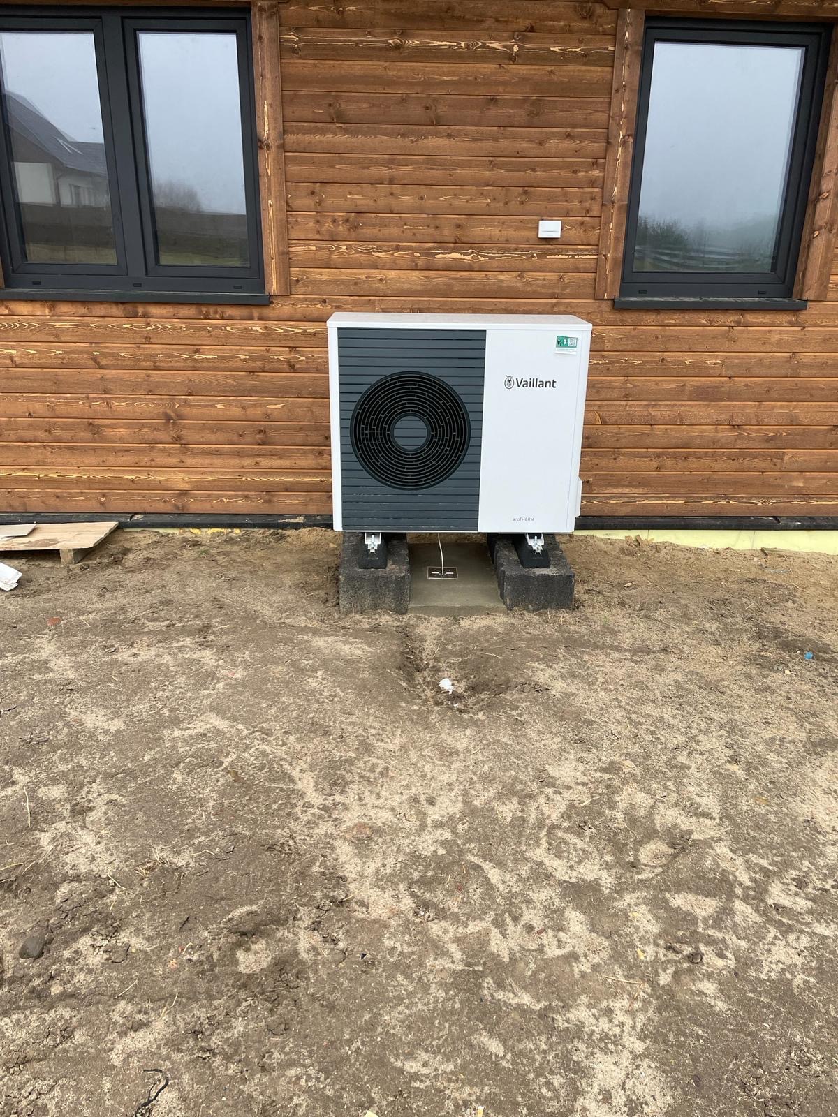 Biała pompa ciepła Vaillant aroTHERM split zamontowana na betonowych podstawach przy drewnianej elewacji domu z czarnymi oknami, widoczne ślady prac ziemnych.